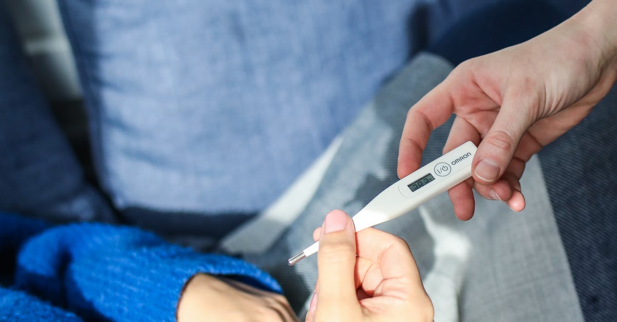 A woman checks her temperature using a digital thermometer while resting on a couch at home.