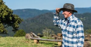 A smiling farmer in plaid flannel enjoys a sunny day on a lush green hillside.