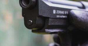 A detailed close-up of a gloved hand holding and aiming a semi-automatic pistol outdoors.