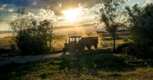 A tractor moves through a rural landscape at sunrise, with sunrays filtering through trees and creating a scenic view.